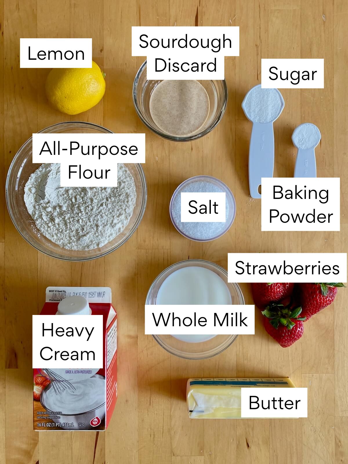 The ingredients to make sourdough strawberry shortcake. Each ingredient is labeled with text. They include sourdough discard, lemon, sugar, all-purpose flour, salt, baking powder, heavy cream, whole milk, strawberries, and butter.