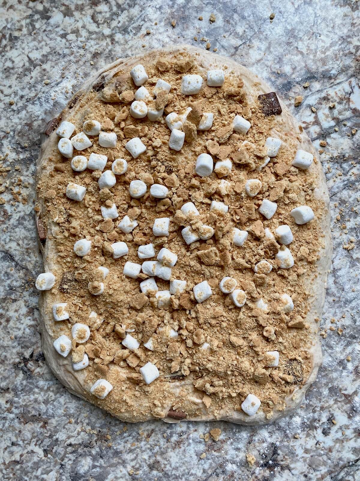 Crushed graham crackers and mini marshmallows spread out on top of s'mores sourdough bread dough before being rolled up.