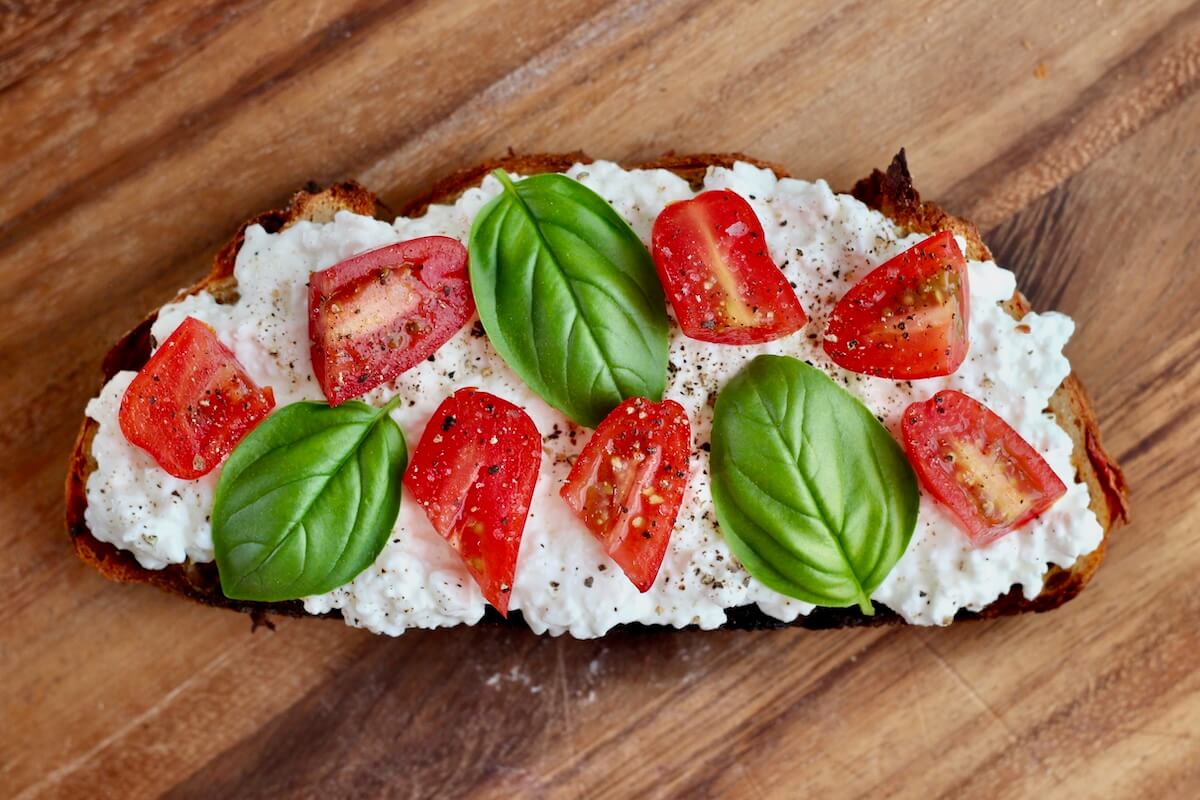 A piece of sourdough cottage cheese toast topped with tomatoes, basil, salt, and pepper.