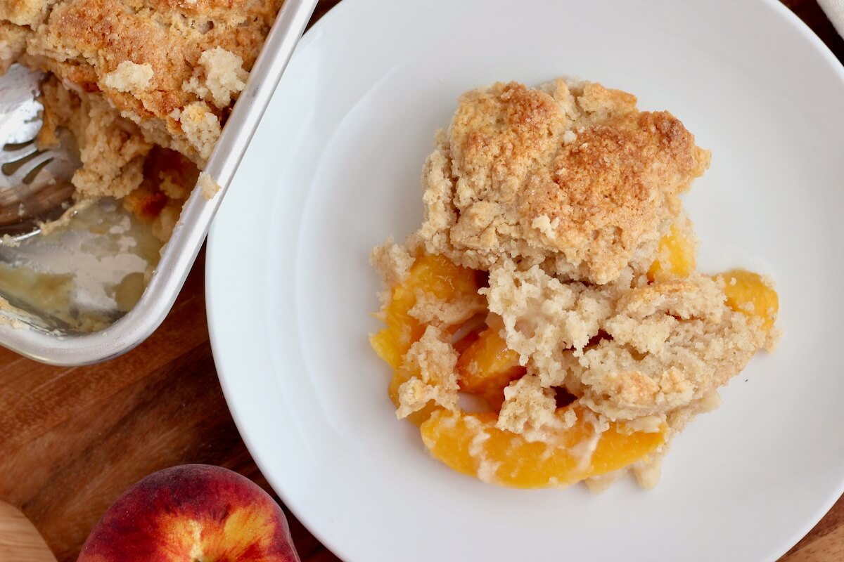 A serving of sourdough peach cobbler on a small plate next to a baking dish full of more cobbler.