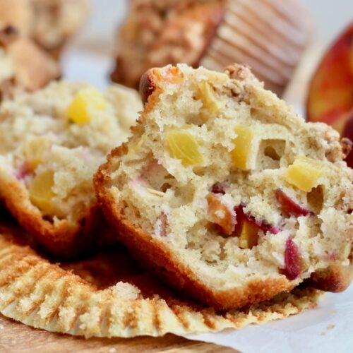 A sourdough peach muffins split in half on a paper wrapper. There are more muffins and a whole peach out of focus in the background.