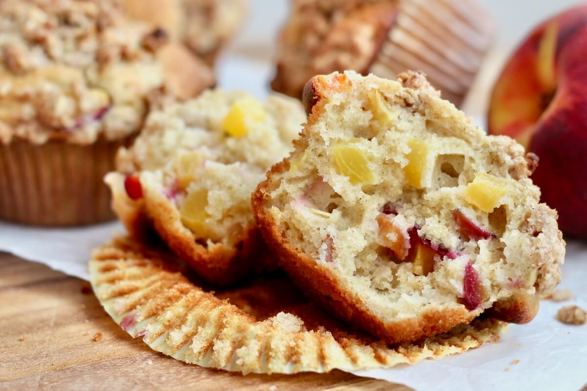 A sourdough peach muffins split in half on a paper wrapper. There are more muffins and a whole peach out of focus in the background.