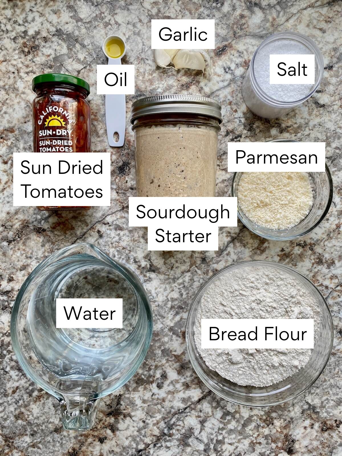 The ingredients to make sun dried tomato sourdough bread. Each ingredient is labeled with text. They include bread flour, water, sourdough starter, salt, sun dried tomatoes, parmesan cheese, garlic, and oil.