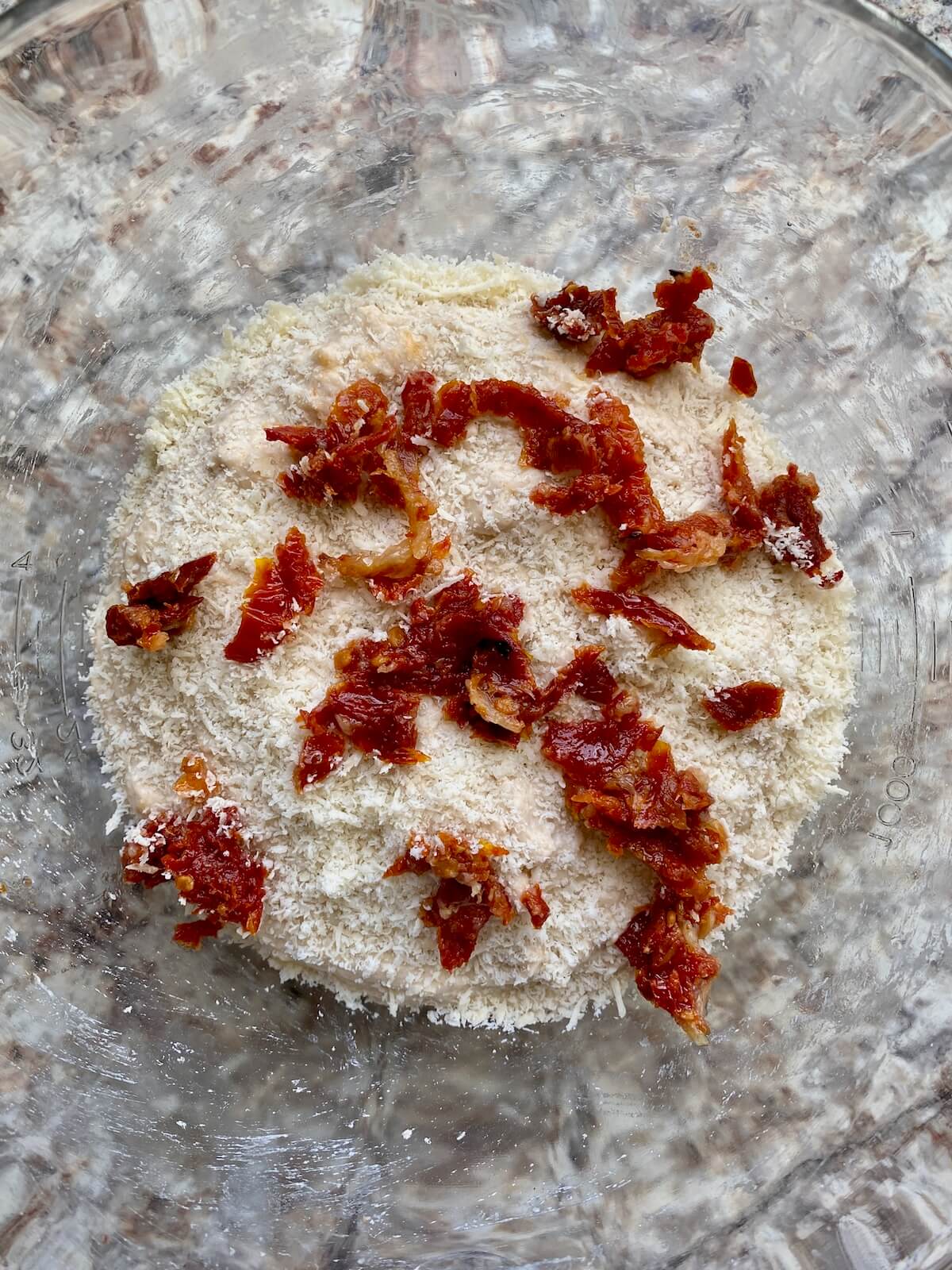 Sourdough bread dough topped with sun dried tomatoes, roasted garlic, and grated parmesan cheese in a bowl.