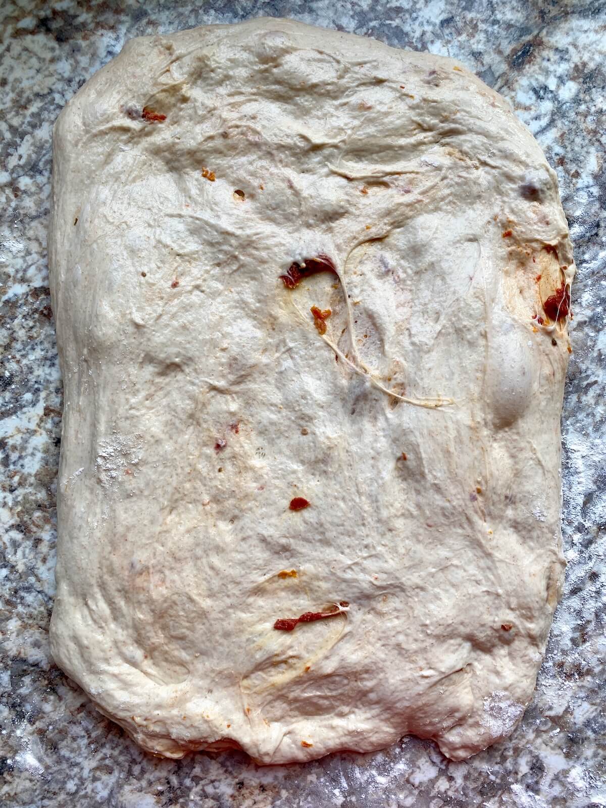 Sun dried tomato and roasted garlic sourdough bread dough stretched into a rectangle on a floured counter.