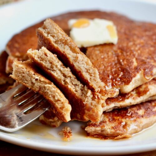 Three whole wheat sourdough pancakes on a plate topped with butter and maple syrup. A bite of all three pancakes has been cut out and are on a fork situated on the plate.