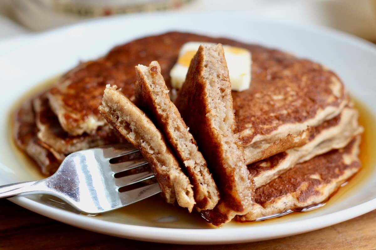 A stack of three sourdough banana pancakes. A fork is resting on the plate with three pieces of pancake on it.