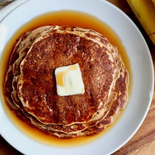 A stack of sourdough banana pancakes with a pat of butter and a generous amount of maple syrup pooling around them.