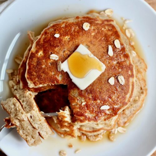 A stack of sourdough oatmeal pancakes with a pat of butter and maple syrup on top. There is a triangular-shaped bite taken out with the cut out pieces on a fork laying against the plate.