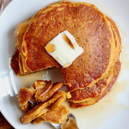 A stack of sourdough pumpkin pancakes with a pat of butter and maple syrup. There is a forkful of pancake resting on the plate.