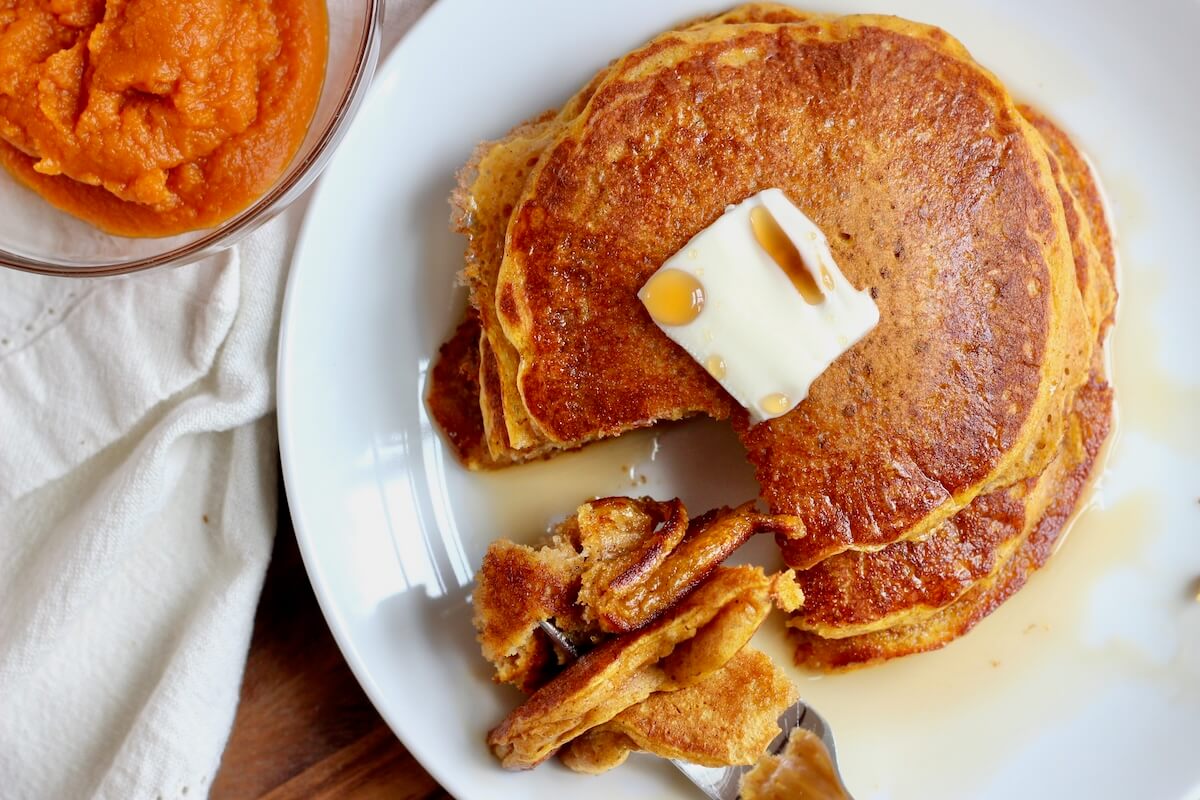 A stack of sourdough pumpkin pancakes with a pat of butter and maple syrup. There is a forkful of pancake resting on the plate and a small bowl of pumpkin puree next to the plate.