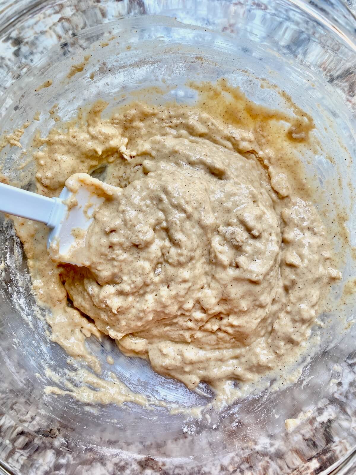 The wet and dry ingredients combined in a glass bowl. There is a rubber spatula sticking out of the bowl to the left.