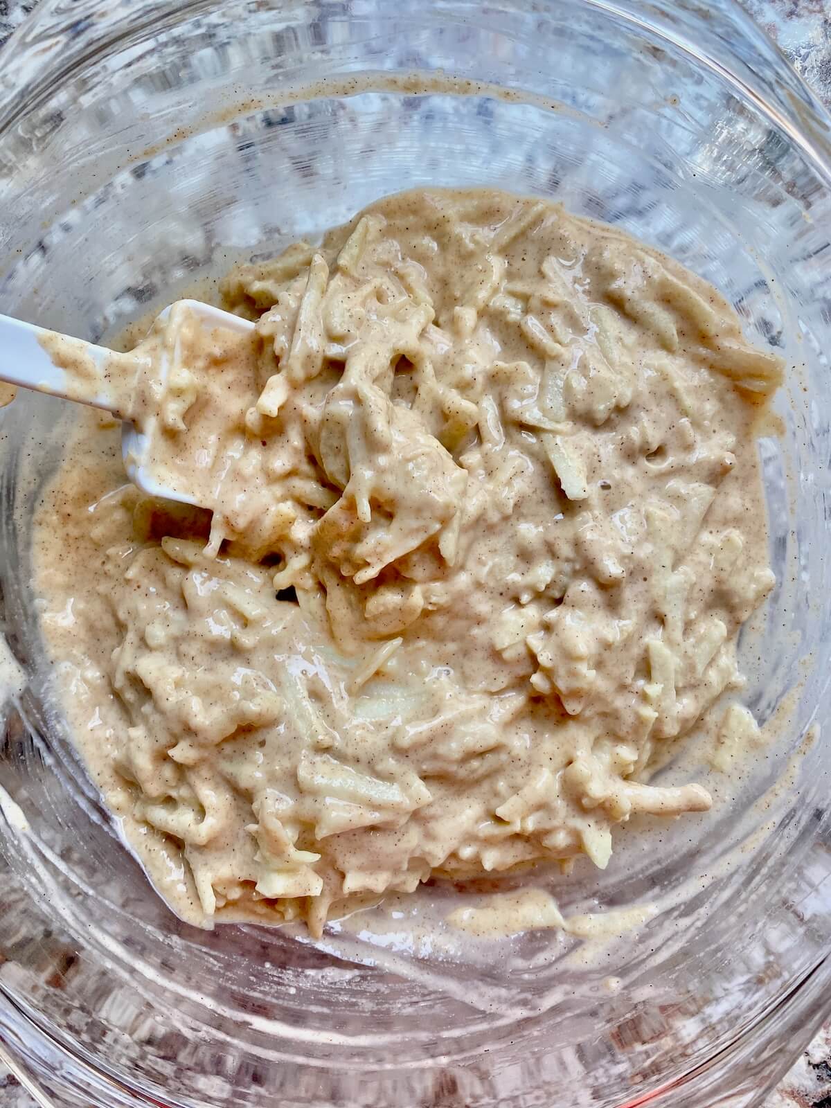 The sourdough apple fritter batter in a glass bowl with a rubber spatula sticking out to the left.