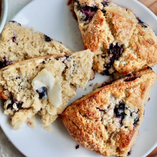 Three blueberry sourdough scones on a plate. One of the scones is split in half with butter on it.