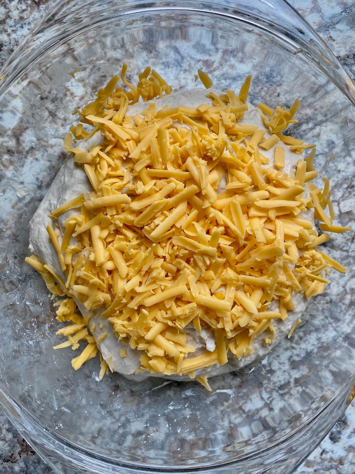 A sourdough bread dough topped with shredded cheddar cheese in a glass bowl.
