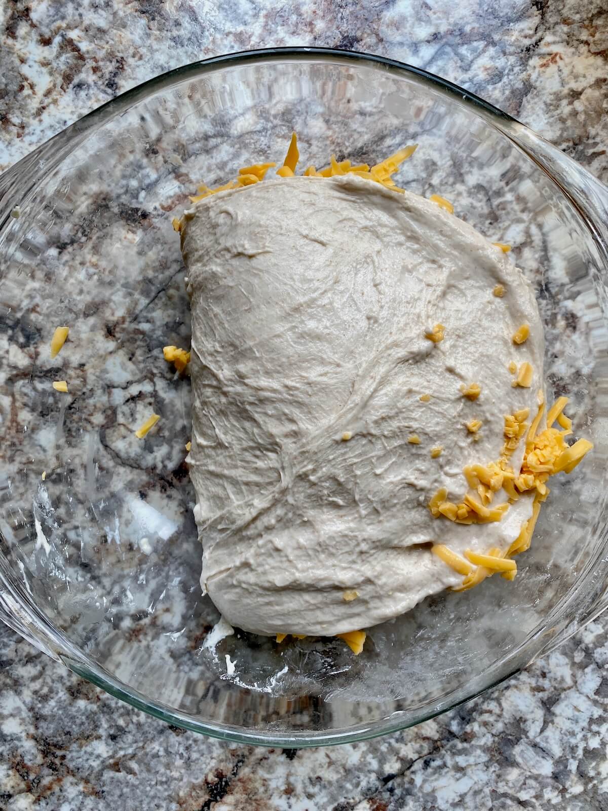 Sourdough bread dough with cheddar cheese folded over itself in a glass bowl.