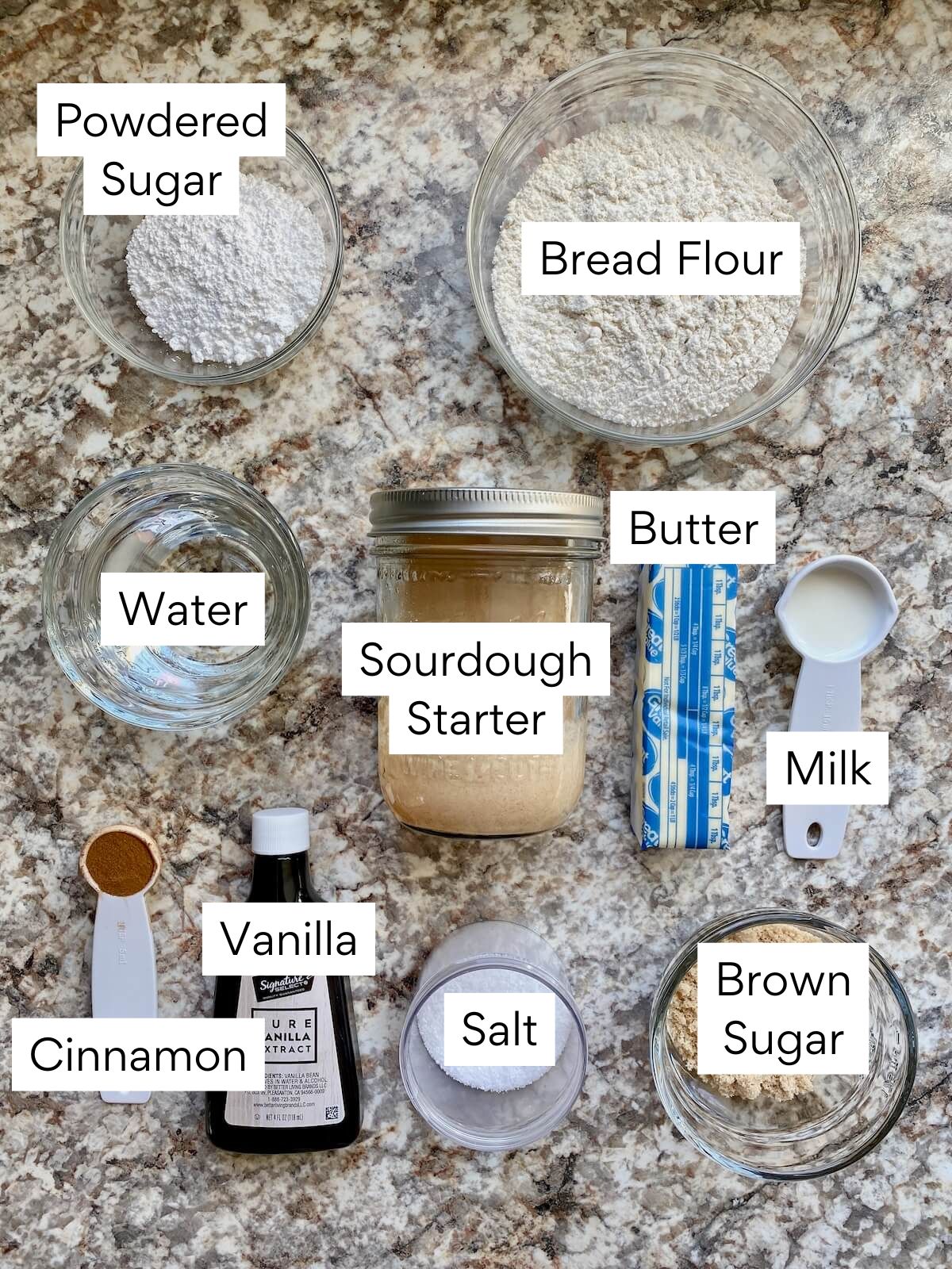 The ingredients to make cinnamon roll sourdough focaccia. Each ingredient is labeled with text. They include bread flour, water, sourdough starter, butter, milk, cinnamon, vanilla, salt, brown sugar, and powdered sugar.