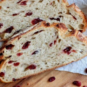 Slices of cranberry orange sourdough bread on a piece of parchment paper. Around the bread slices are a few dried cranberries.