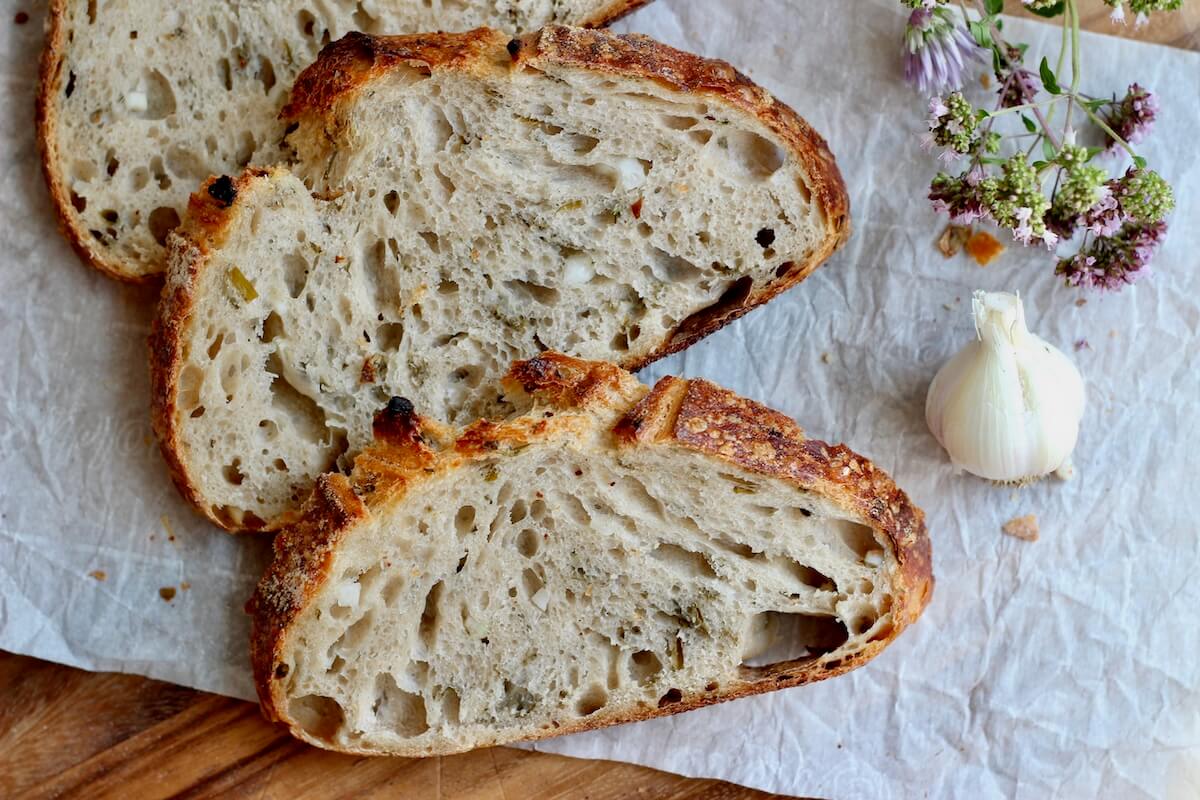 Three slices of garlic sourdough bread with fresh herbs on a piece of wrinkled parchment paper.