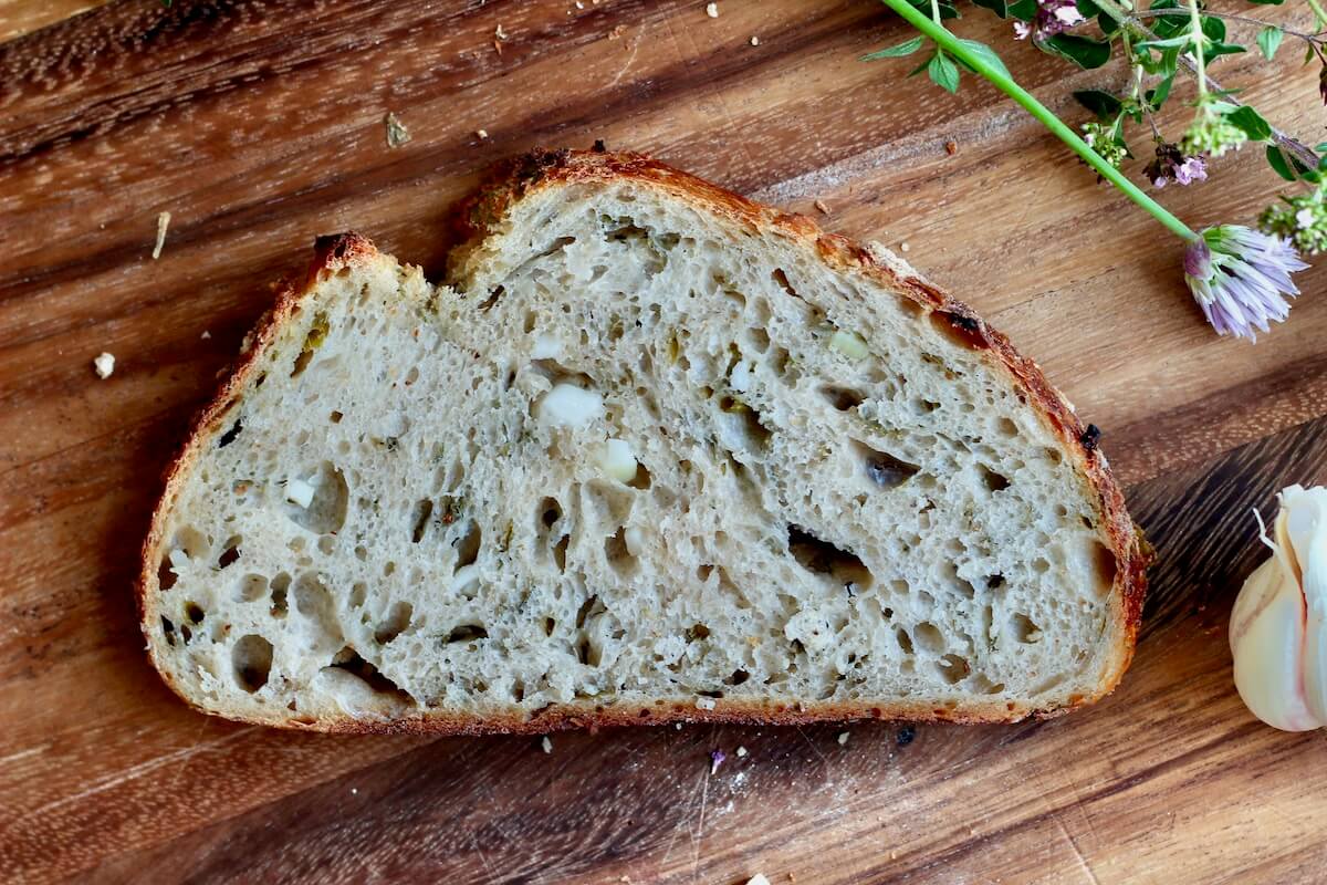 A slice of garlic sourdough bread on a wooden cutting board. A head of garlic and fresh herbs are laying on the board around the bread.