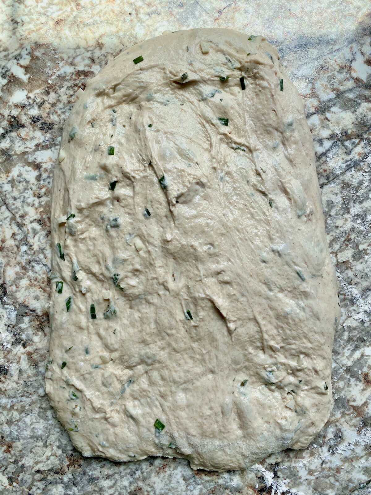 Fermented garlic herb sourdough bread dough stretched out into a rectangle on a floured counter.