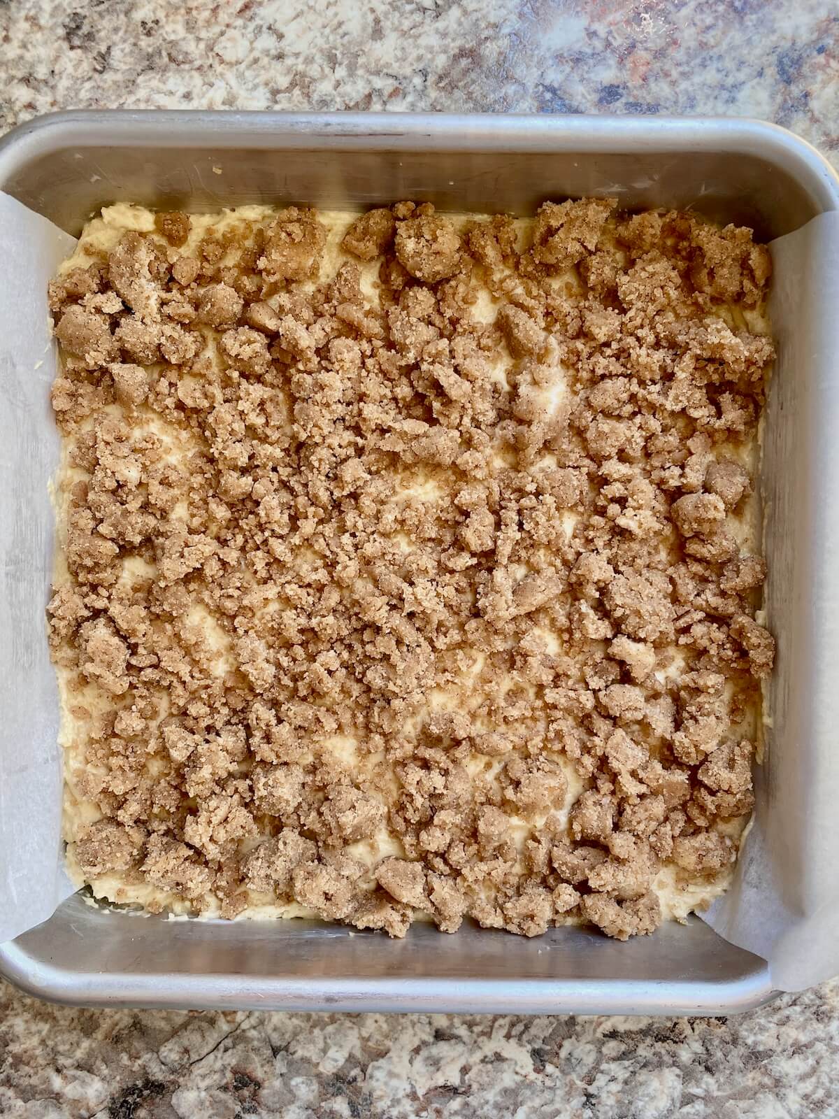 A square aluminum cake pan lined with parchment paper with one layer of cake batter topped with a layer of crumble filling.