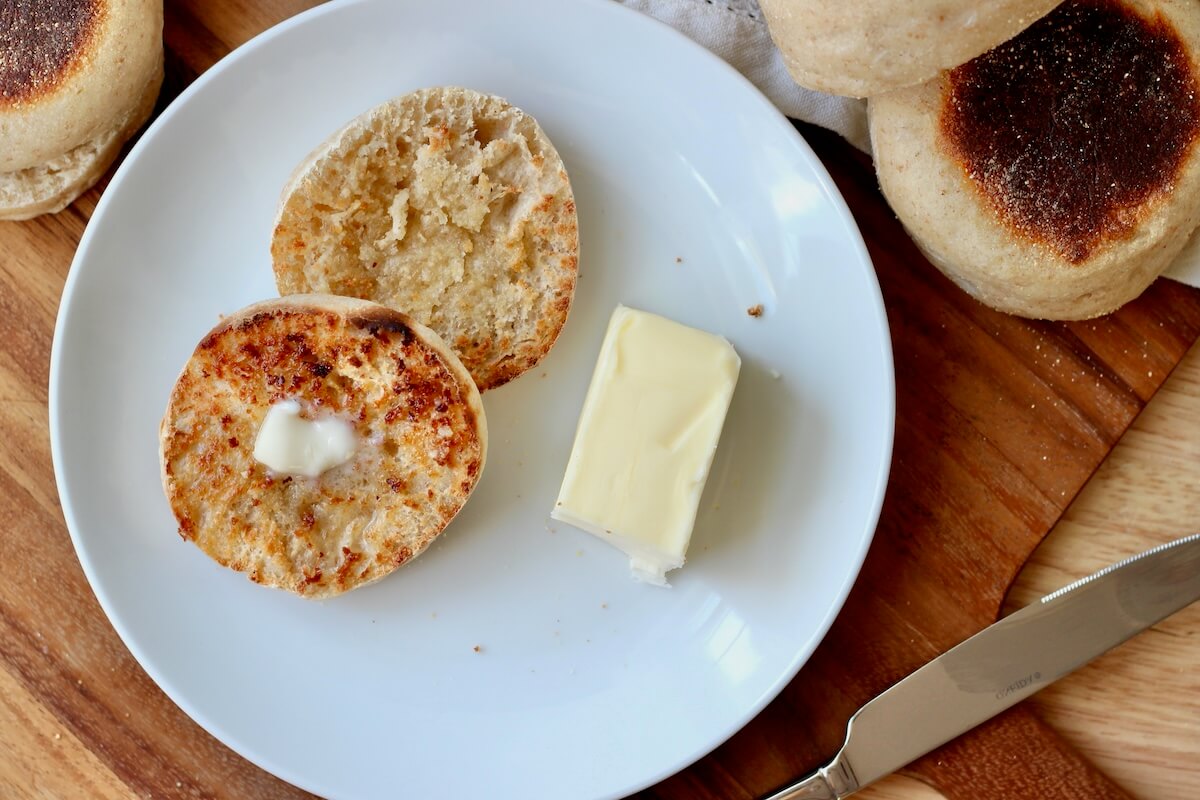 A toasted and buttered sourdough English muffin on a small white plate. There is half a stick of butter on the plate next to the English muffin. Several more English muffins and a butter knife are situated around the plate.
