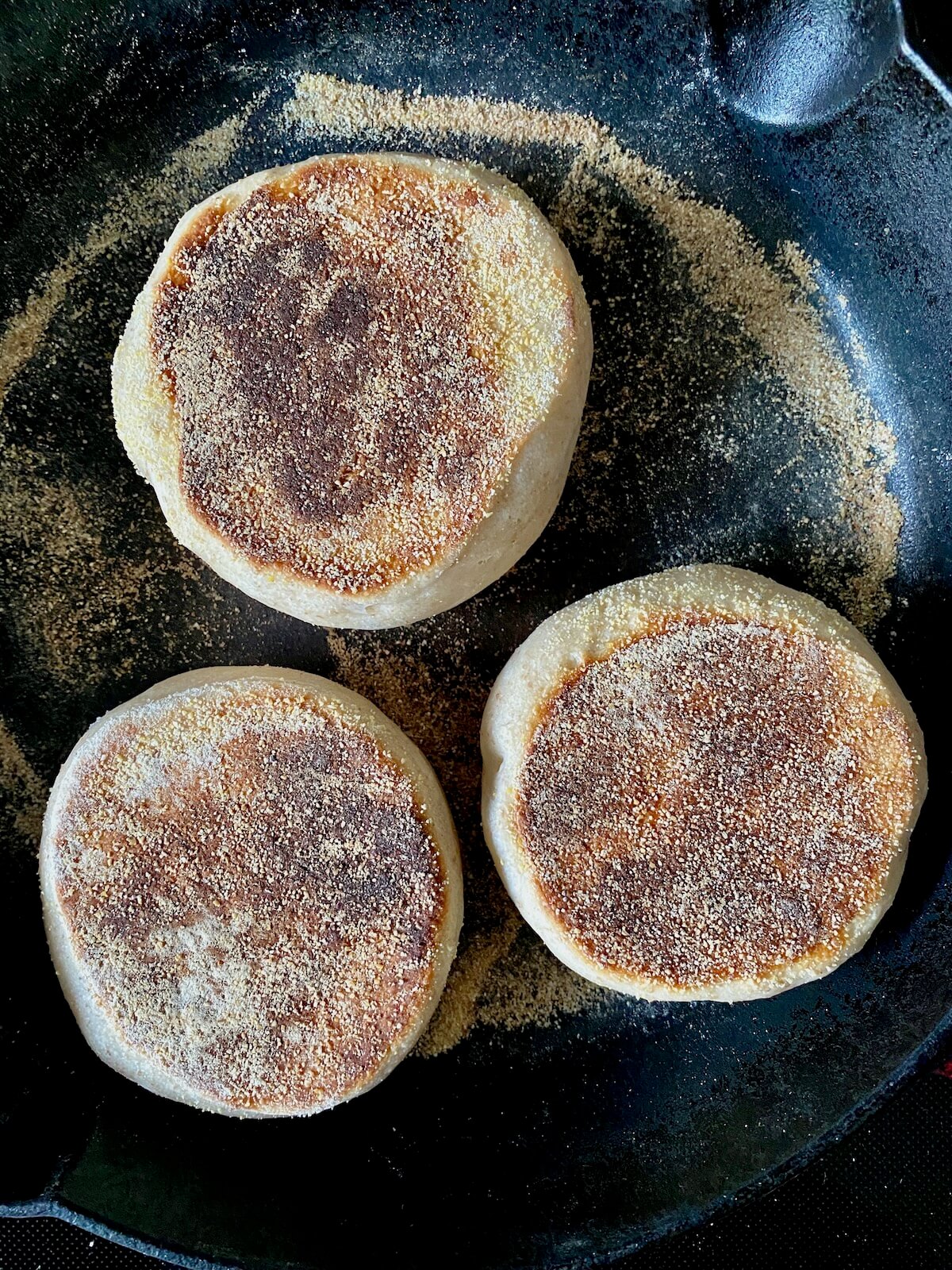 Three partially-cooked sourdough English muffins cooking in a cornmeal-dusted cast iron skillet.