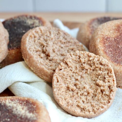 Several whole wheat sourdough English muffins on a white linen cloth. One of the muffins is split open to show the nooks and crannies.