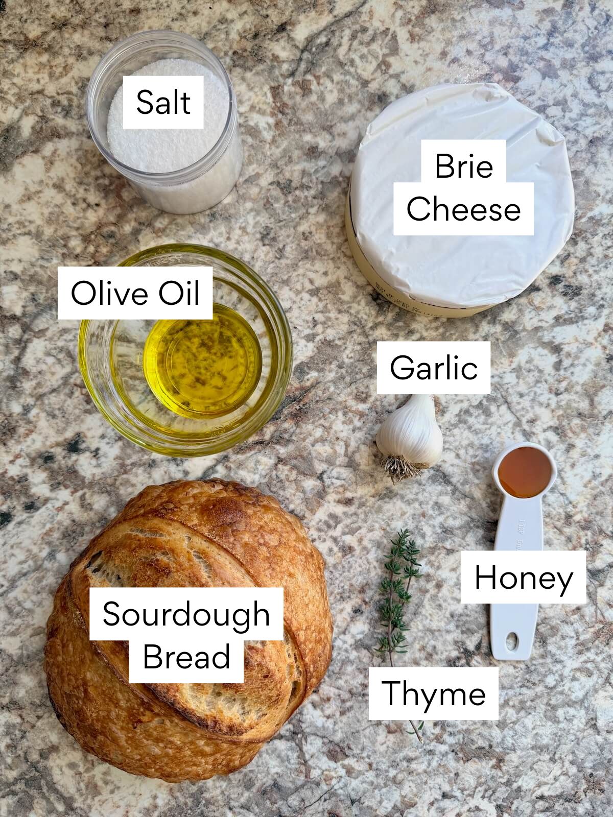 The ingredients to make baked brie in sourdough. Each ingredient is labeled with text. They include brie cheese, sourdough bread, olive oil, garlic, honey, thyme, and salt.