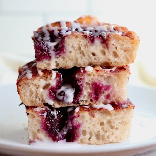 Three pieces of sourdough blueberry focaccia stacked on top of each other on a small, white plate.