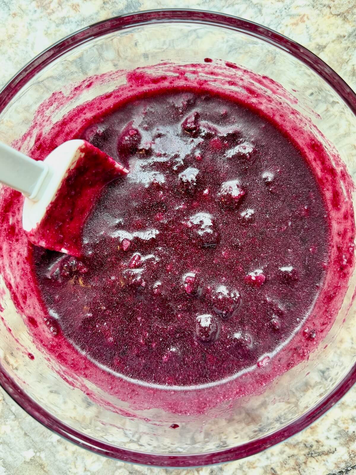 Homemade blueberry sauce in a glass bowl with a rubber spatula sticking out to the left.