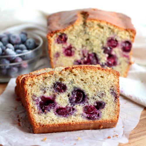 A few slices of sourdough blueberry bread set on a piece of parchment paper. The other half of the loaf and a small bowl of blueberries are out of focus in the background.