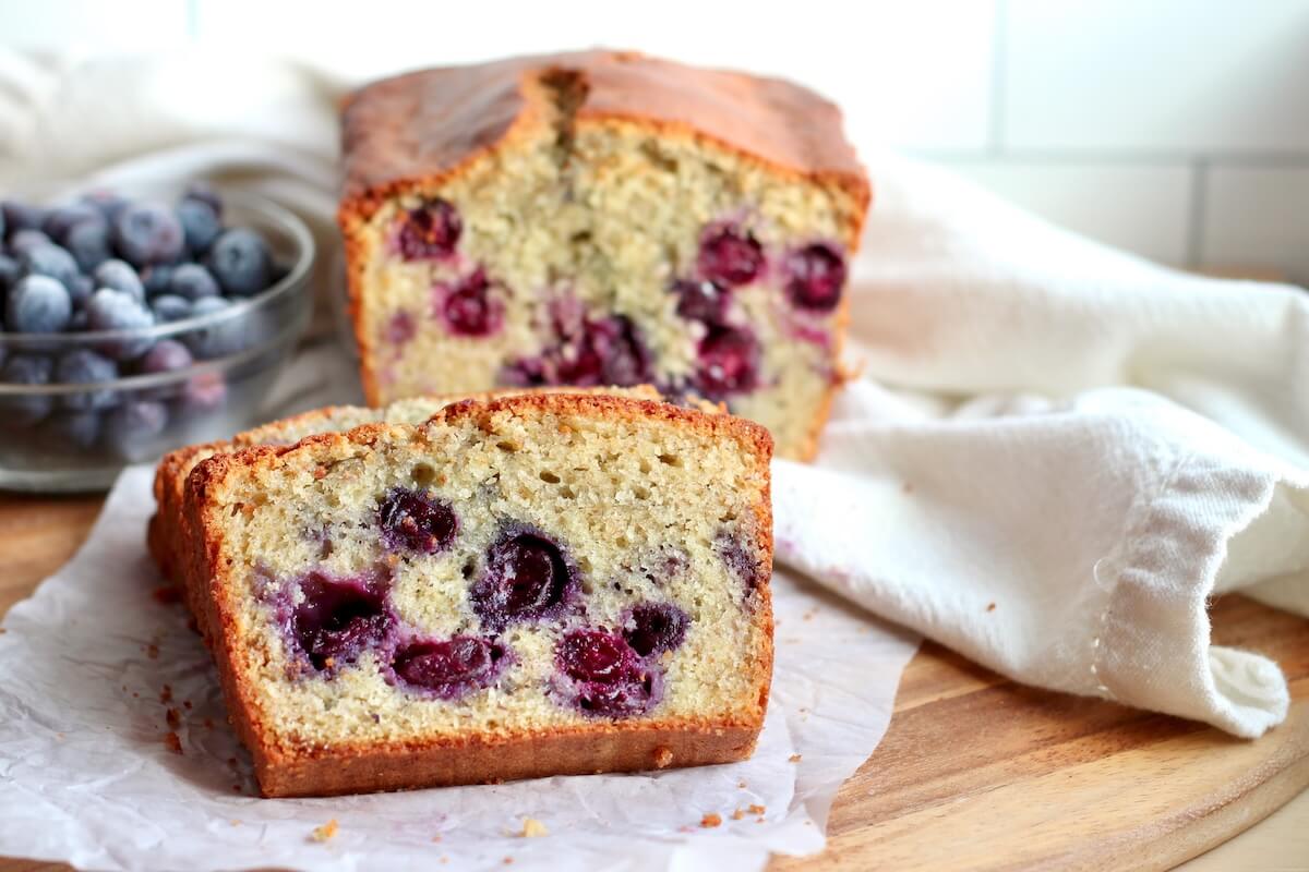 A few slices of sourdough blueberry bread set on a piece of parchment paper. The other half of the loaf and a small bowl of blueberries are out of focus in the background.