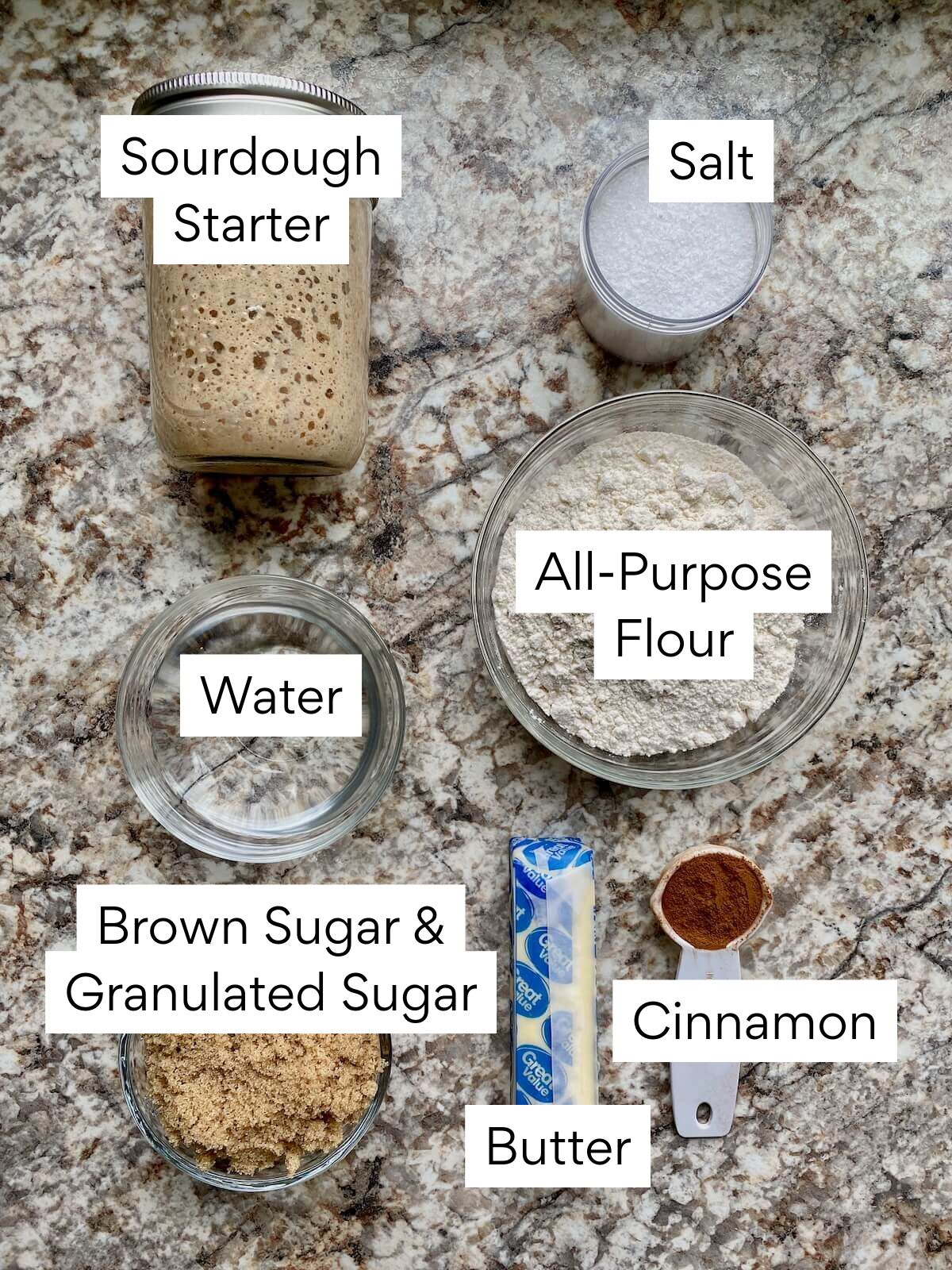 The ingredients to make sourdough monkey bread. Each ingredient is labeled with text. They include sourdough starter, salt, water, all-purpose flour, brown sugar, granulated sugar, butter, and cinnamon.