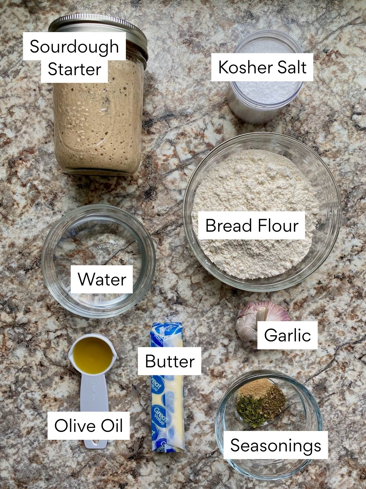The ingredients to make sourdough pull apart garlic bread. Each ingredient is labeled with text. They include sourdough starter, kosher salt, water, bread flour, olive oil, butter, garlic, and seasonings.