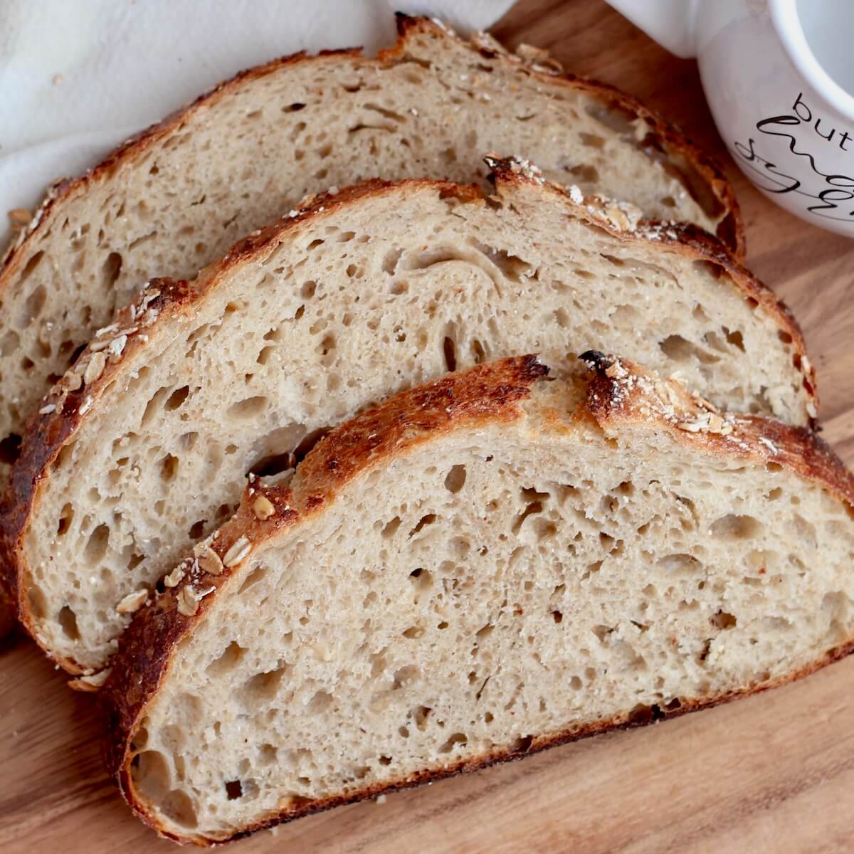 Three slices of maple oat sourdough bread on a cutting board. On the cutting board next to the bread is a white cloth napkin and a ceramic cup that says "but first, maple syrup."