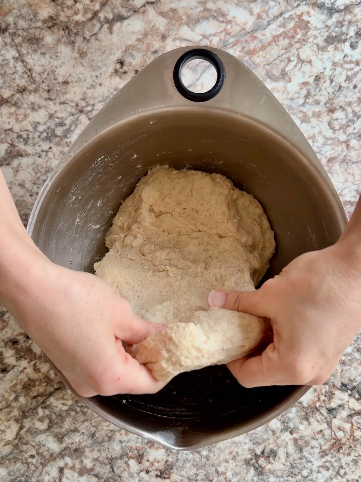 The dough being stretched upwards out of a stainless steel bowl by two hands.
