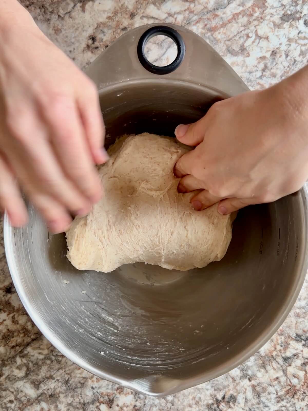 The stretched dough being folded over itself in a stainless steel bowl by two hands.