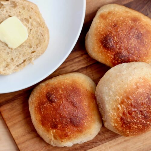 Three sourdough potato rolls on a cutting board. Out of focus to the left of the rolls is a split potato roll with butter on a plate.
