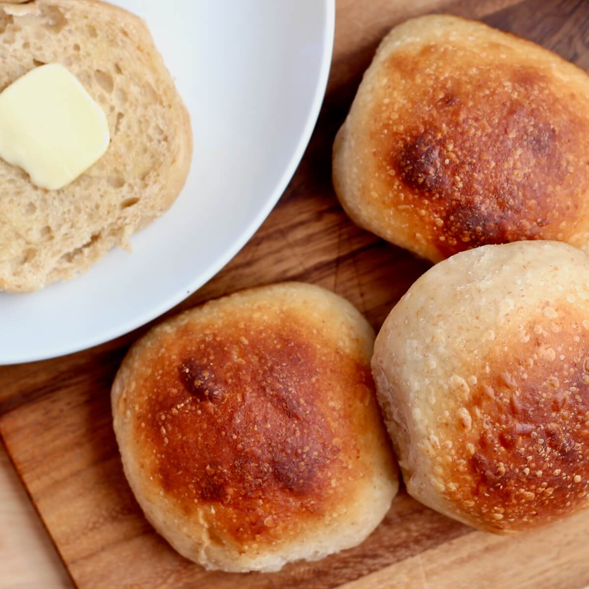 Three sourdough potato rolls on a cutting board. Out of focus to the left of the rolls is a split potato roll with butter on a plate.