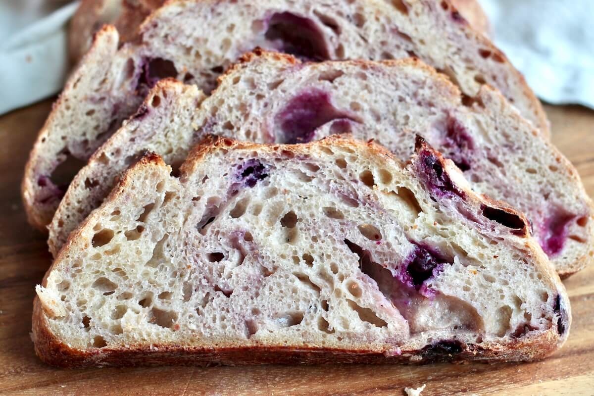 A sliced loaf of blueberry sourdough bread on a wooden cutting board.