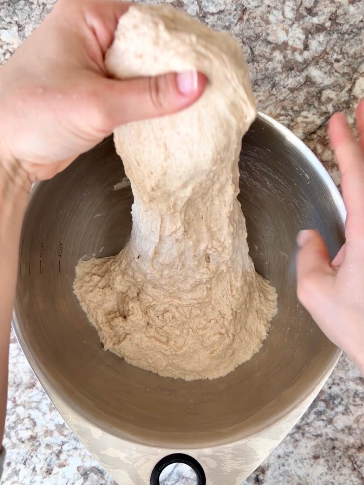 Two hands stretching dough upwards out of a stainless steel bowl.