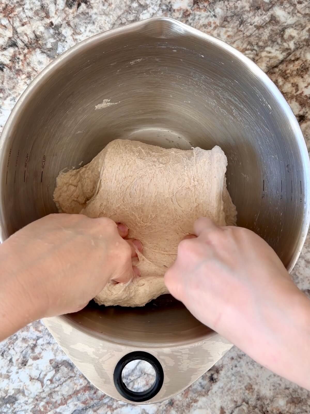 Two hands folding dough over itself in a stainless steel bowl.