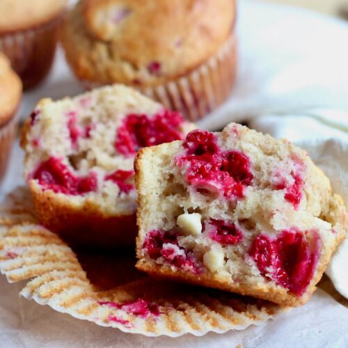 A raspberry white chocolate sourdough muffin split in half on a paper liner with several other muffins out of focus in the background.