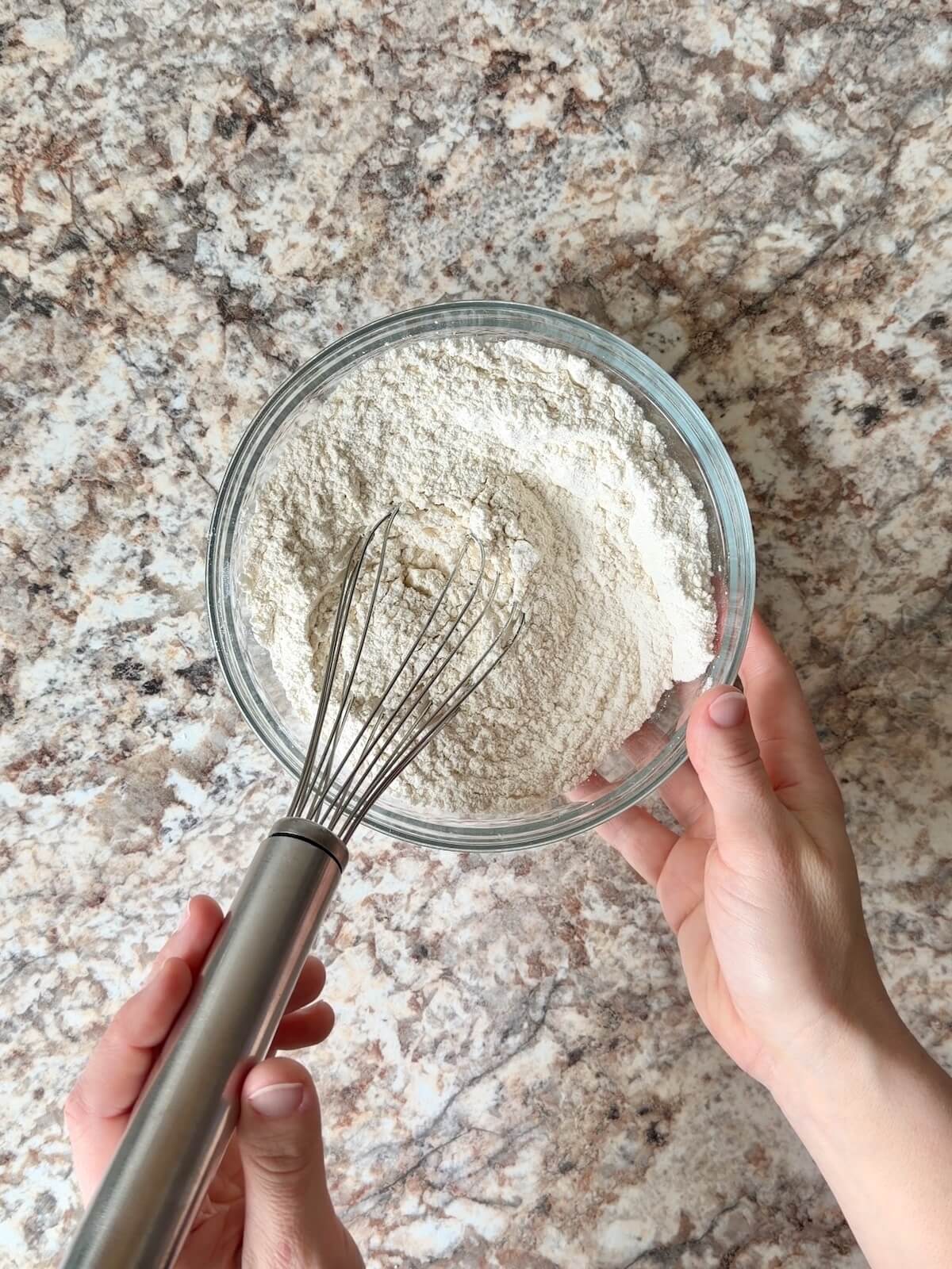 Two hands whisking a bowl of flour and other dry ingredients together in a glass bowl.