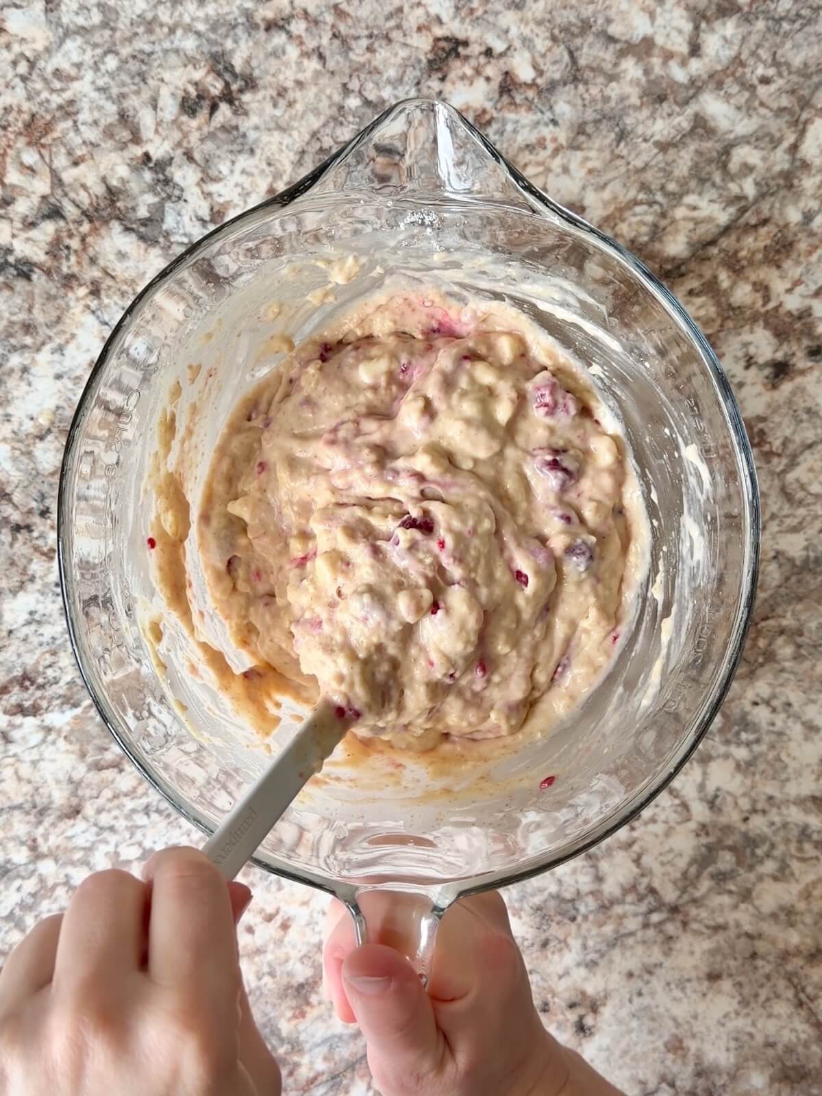 White chocolate raspberry sourdough muffin batter being mixed with a rubber spatula in a glass bowl.