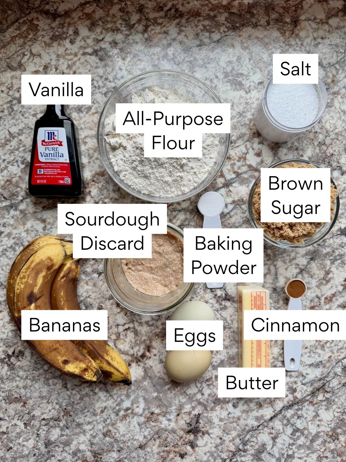 The ingredients to make sourdough banana bread. Each ingredient is labeled with text. They include vanilla, all-purpose flour, salt, sourdough discard, baking powder, brown sugar, bananas, eggs, butter, and cinnamon.