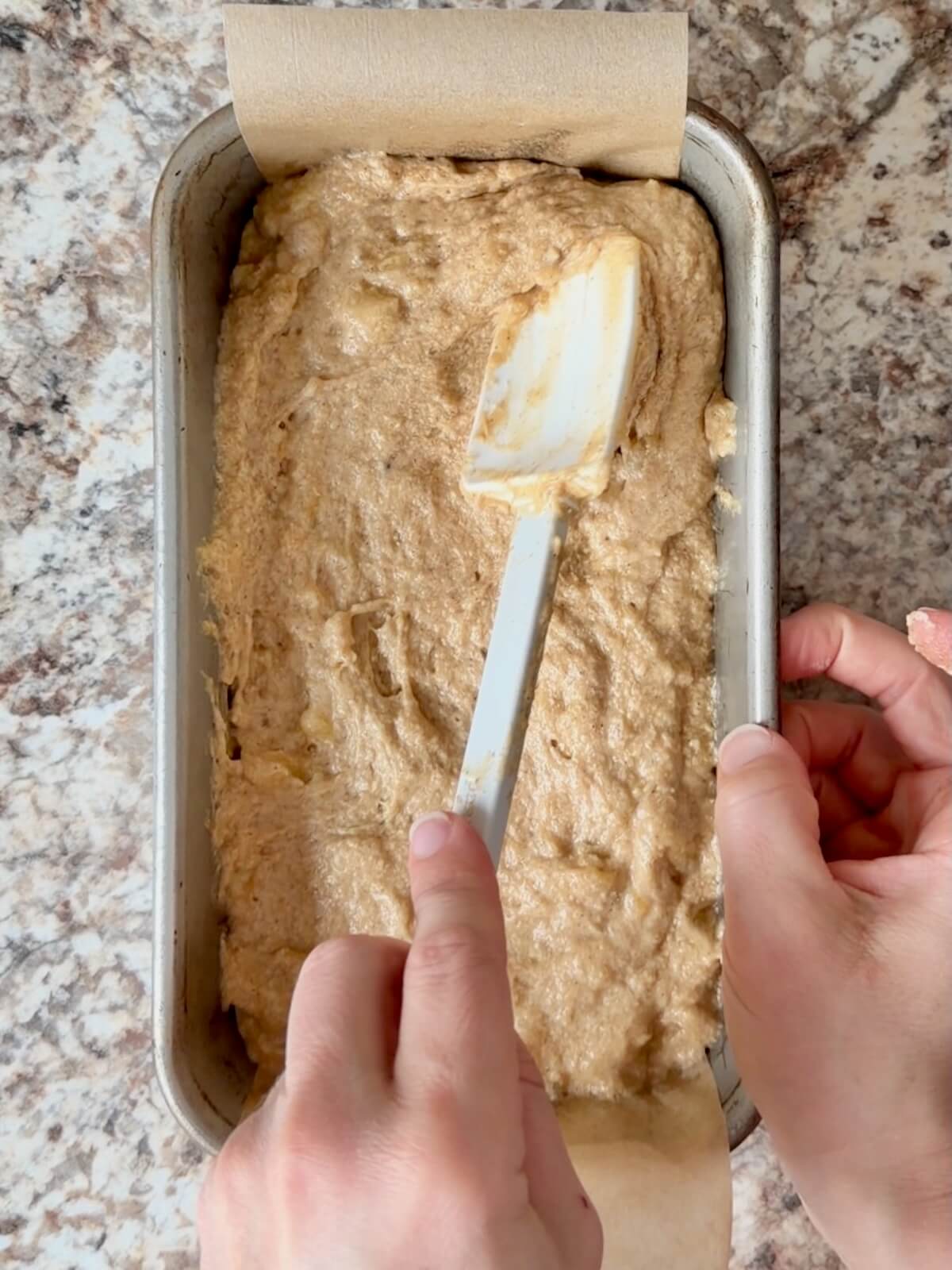 Sourdough banana bread batter being spread into a parchment lined loaf pan using a rubber spatula.