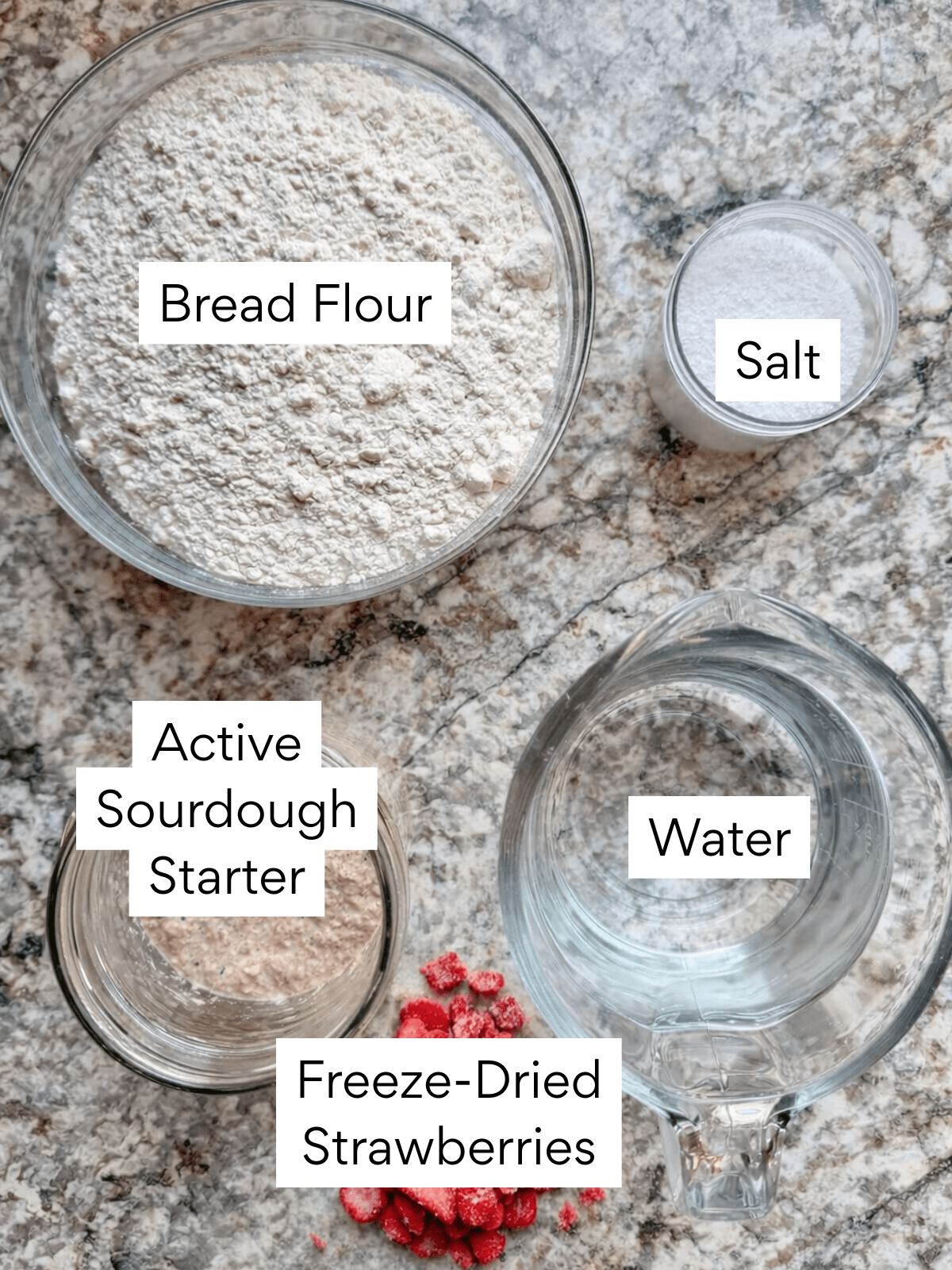 The ingredients to make strawberry sourdough bread. Each ingredient is labeled with text. They include bread flour, salt, active sourdough starter, water, and freeze-dried strawberries.